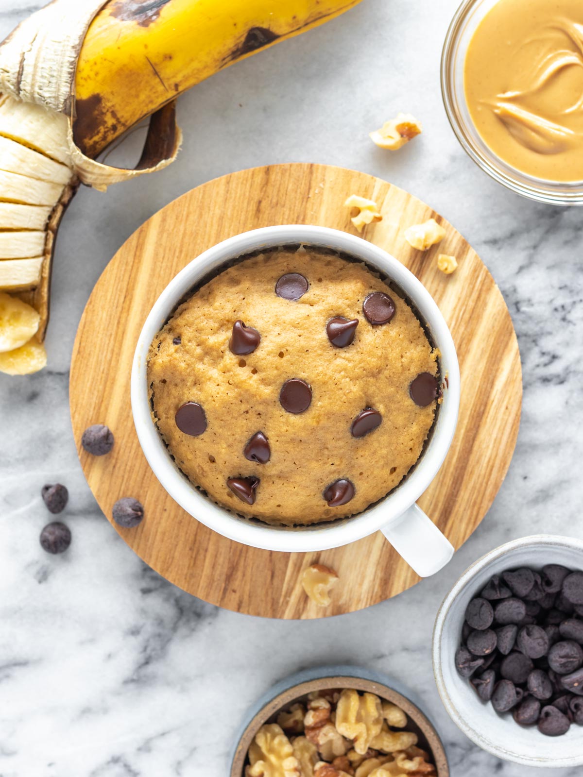bolo de caneca de banana visto de cima, com uma banana, pasta de amendoim, nozes picadas e chocolate em gotas ao redor dele