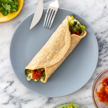 panqueca de aveia recheada com homus, brócolis, ervilha e tomate, servida em um prato azul