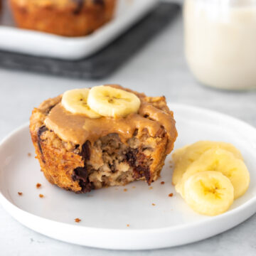 Muffin de banana com aveia servido com uma cobertura de pasta de amendoim natural e duas rodelas de banana por cima. O muffin está mordido, mostrando sua textura única no meio, com gotas de chocolate derretidas.