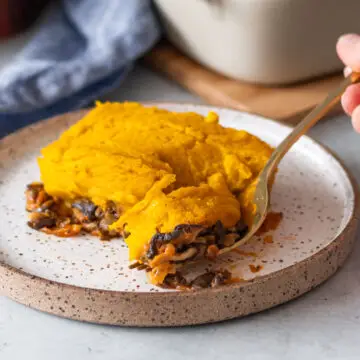 pedaço de escondidinho de cogumelos com cobertura de purê de abóbora servido em um prato, com um pedaço retirado em cima de um garfo