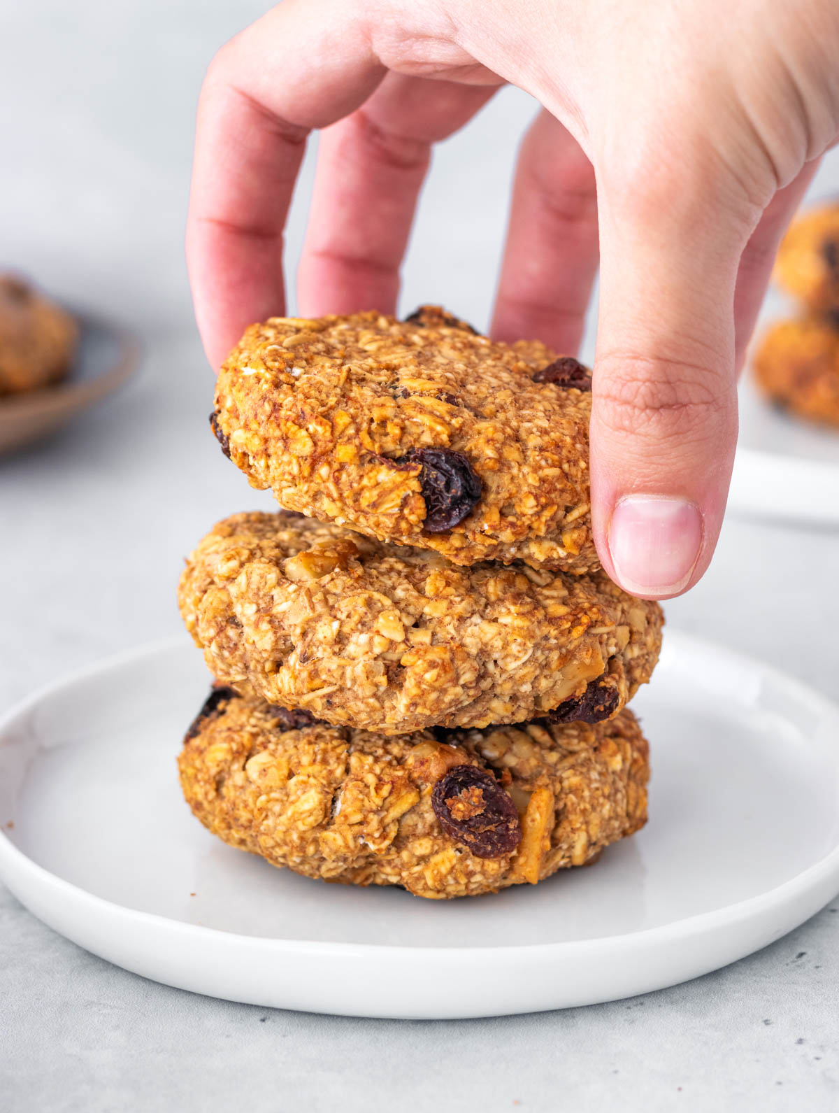 3 cookies de aveia e banana empilhados em um prato, com uma mão pegando o de cima
