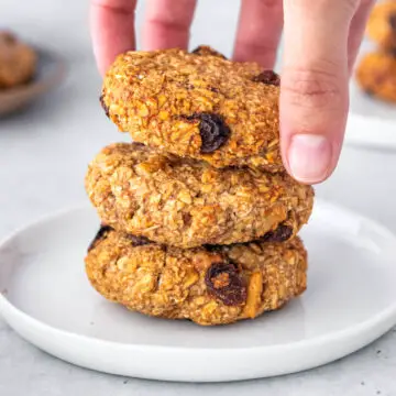 3 cookies de aveia com banana empilhados em um prato, com uma mão pegando o de cima