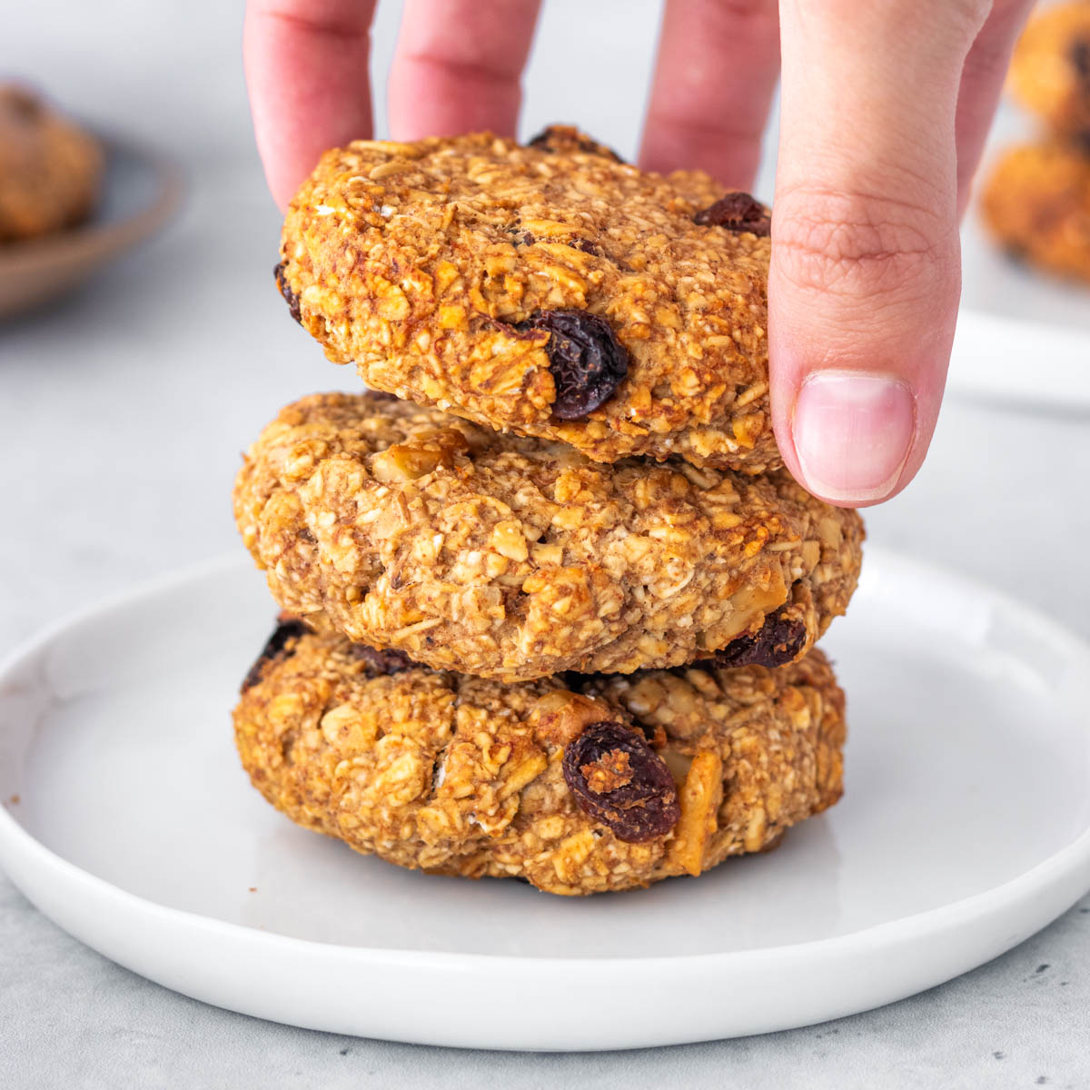 3 cookies de aveia com banana empilhados em um prato, com uma mão pegando o de cima