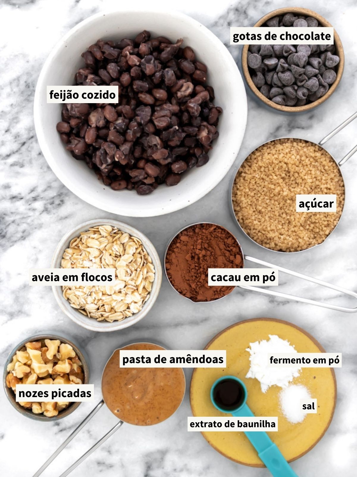 ingredientes para fazer brownie de feijão, incluindo aveia em flocos grossos, cacau em pó, açúcar demerara, sal, feijão cozido (feijão preto), pasta de amêndoas natural, extrato de baunilha, bicarbonato de sódio, nozes picadas (nozes-chilenas) e gotas de chocolate