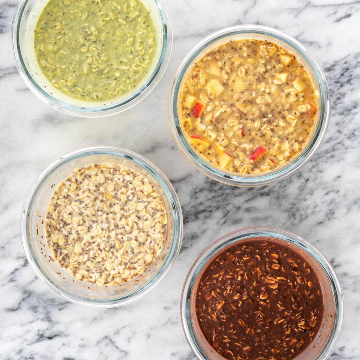 quatro potes com diferentes sabores de overnight oats (matcha, maçã com canela, básico e banana com chocolate) sem tampa em cima de uma bancada