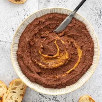 espátula sendo inserida em pote com pasta de cacau e amendoim feita com resíduo do leite vegetal servida em um prato com mais pasta de amendoim por cima, mostrando sua textura cremosa