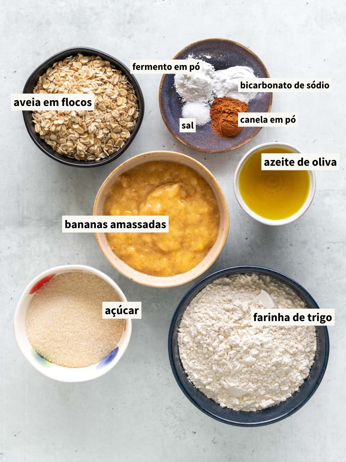 ingredientes para fazer bolo de banana com aveia, incluindo purê de banana madura, açúcar, canela em pó, sal, água filtrada, azeite de oliva, extrato de baunilha, farinha de trigo, fermento químico em pó, bicarbonato de sódio e aveia em flocos grossos