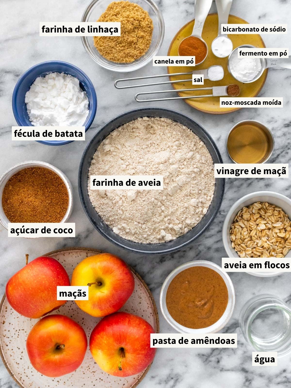 ingredientes para fazer bolo de maçã com aveia, incluindo farinha de aveia fina, maçã fresca, pasta de amêndoas, açúcar, farinha de linhaça, água filtrada, canela em pó, sal, noz-moscada em pó, vinagre de maçã, fécula de batata, fermento químico em pó e bicarbonato de sódio