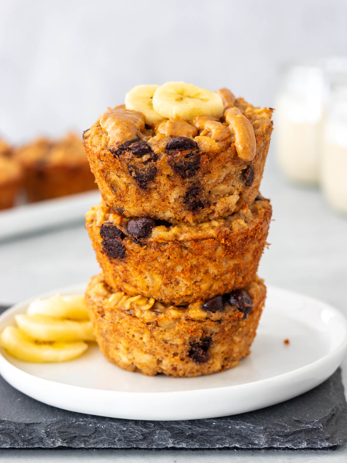 três muffins de banana com aveia e gotas de chocolate empilhados em um prato, com cobertura de pasta de amendoim e rodelas de banana