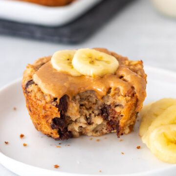 muffin de banana com aveia servido em um prato, coberto com pasta de amendoim e rodelas de banana, com uma mordida mostrando sua textura úmida com porções de chocolate derretido