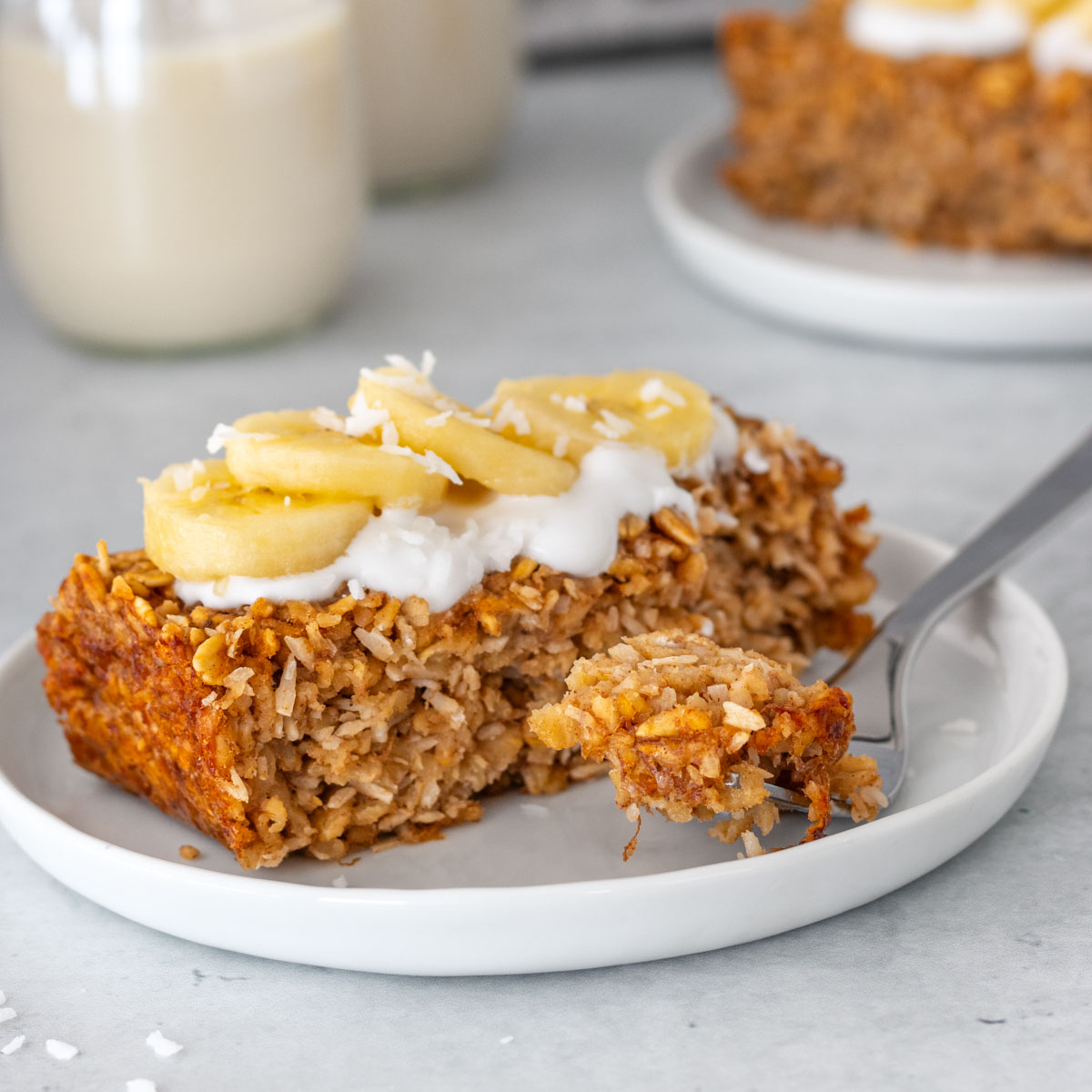fatia de bolo de mingau de aveia assado com coco e banana servido em um prato com iogurte de coco, rodelas de banana fresca e coco ralado