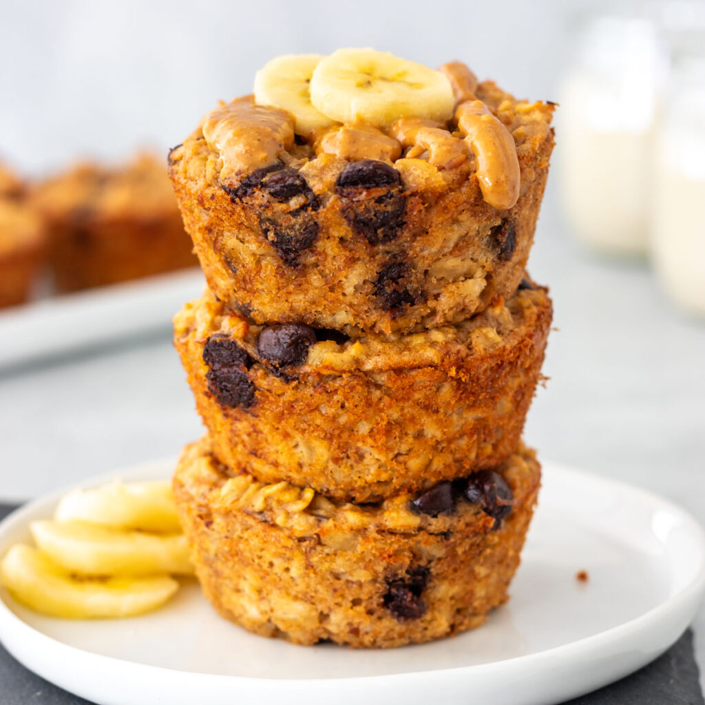 três muffins de banana com aveia e gotas de chocolate empilhados em um prato, com cobertura de pasta de amendoim e rodelas de banana